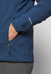 Main d'une personne dans la poche d'une veste WINDWALL bleue, montrant le détail de la fermeture éclair et le poignet élastique, avec un pantalon gris visible sous la veste.