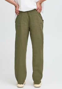 Person wearing olive green pants with back pockets and white sneakers, standing with hands in pockets against a plain background.