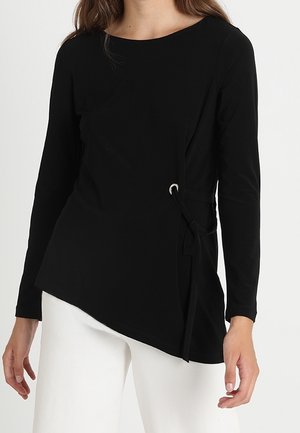 Vrouw die een zwart shirt met lange mouwen draagt, met een asymmetrische zoom en een detail met een strik aan de zijkant, gecombineerd met een witte broek.