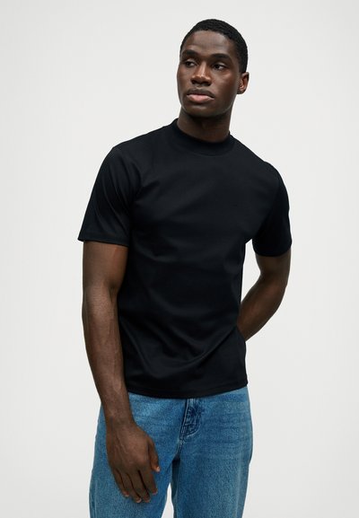 Νεαρός άνδρας φορώντας ένα εφαρμοστό μαύρο κοντομάνικο πουκάμισο και μπλε τζιν, κοιτάζοντας προς το πλάι με φόντο μια απλή φωτισμένη επιφάνεια.