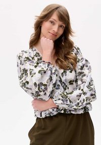 Blumenbluse mit luftigen Ärmeln, versehen mit einem weißen Hintergrund und grünen sowie blauen Mustern. Braune Hose rundet das Outfit ab.