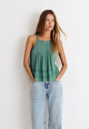 Jeune femme aux longs cheveux bruns portant un haut vert plissé sans manches et un jean bleu clair, debout les mains dans les poches.