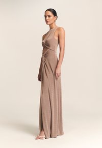 Długa, dopasowana brązowa sukienka z węzłowym wzorem w talii i wysokim dekoltem. Tkanina ma delikatny połysk i miękką fakturę.