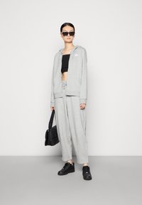 Siva zip-up kapuca s vezalko, črn kratki zgornji del, oversize sivi hlače, črne superge in črna torba; nosi se z sončnimi očali in uhani v obliki obroča.