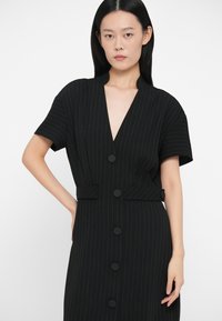 Μαύρο φόρεμα με ρίγες, σε σχήμα V, κοντά μανίκια και εφαρμοστή μέση. Διαθέτει μεγάλα μαύρα κουμπιά και λεία υφή.