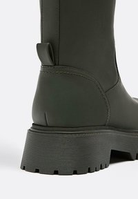 Tmavě zelené gumové kotníkové boty s texturovanou černou platformou, které mají boční švy a zadní poutko pro snadné obouvání.
