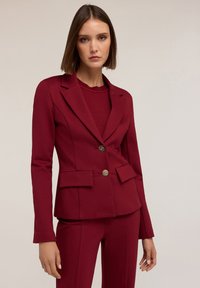 Blazer bordeaux con spalle strutturate, due tasche frontali e bottoni dorati. Tessuto liscio con taglio sartoriale e rever a punta.