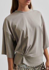 Prevelik taupe T-shirt iz mehkega materiala, z okroglo izreznico in vozlom na pasu. Kratke rokave segajo do komolcev.