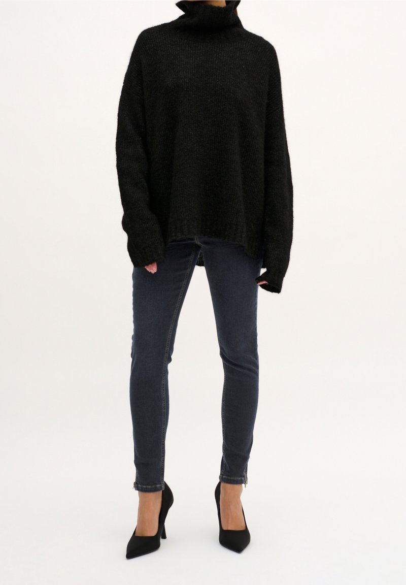 Suéter negro de punto oversized con cuello alto, combinado con jeans ajustados azul oscuro y tacones negros de punta.