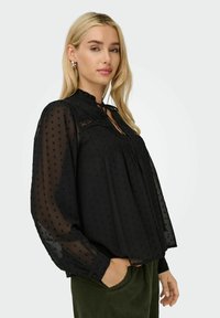 Schwarze Bluse aus durchsichtigem, punktiertem Stoff, mit Rüsche am Hals und gefälteltem Vorderteil. Lange, luftige Ärmel mit geknöpften Manschetten. Strukturiertes Design als Akzent.