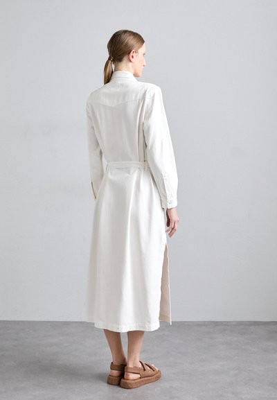 Balta ilga rankovė su diržu iki juosmens, šoninėmis skeltomis ir mygtukais užsegamomis rankovėmis. Ji turi klasikinį apykaklę ir šiek tiek laisvą siluetą.