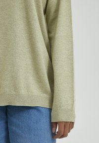 Suéter verde claro con una textura suave y puños acanalados, combinado con unos vaqueros de mezclilla azul, que muestran un ajuste relajado y un diseño sencillo.