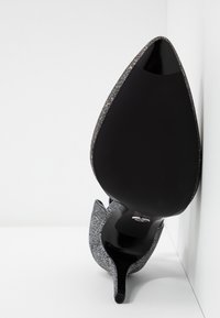 Semelle noire brillante d'un talon aigu avec un dessus argenté scintillant reposant contre un mur d'angle blanc et un sol blanc.