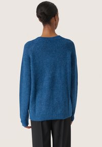 Maglione lavorato a maglia blu con vestibilità morbida, colletto rotondo e polsini a coste, abbinato a pantaloni scuri.