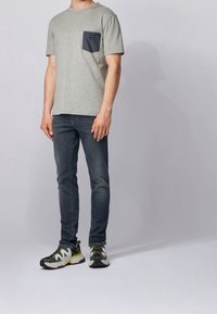 Szary t-shirt z krótkim rękawem z granatową kieszonką na piersi, zestawiony z ciemnymi, obcisłymi dżinsami i wielokolorowymi sneakersami z czarnymi, zielonymi i białymi akcentami.