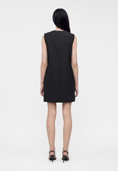 Femme debout, de dos, portant une robe noire sans manches avec une fermeture éclair au dos et des sandales noires à talons hauts sur un fond uni.