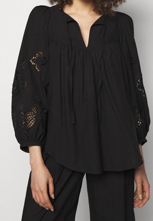Mujer que lleva una blusa negra suelta con patrones de encaje recortados en las mangas abullonadas y pantalones negros de pierna ancha.