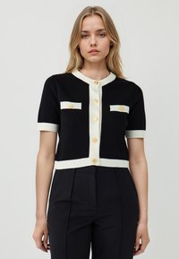 Cardigan nero con maniche corte, bordatura crema e due tasche a toppa. Presenta bottoni dorati e un collo rotondo. Tessuto morbido.