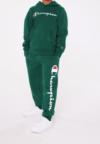 Grön hooded-tröja och sweatpants-set i bomull, med en framficka, vit "Champion" logotyp och vita sneakers.