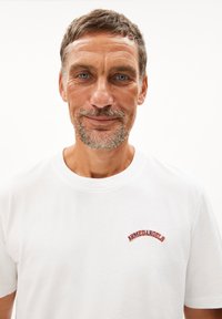 Weißes Baumwoll-T-Shirt mit einem gebogenen, roten gestickten Logo "ARMEDANGELS" auf der Brust. Kurze Ärmel, runder Ausschnitt, lässige Passform.