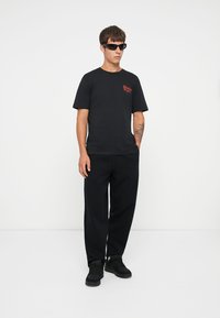 Sort bomuld t-shirt med rød logo på brystet, parret med sorte løstsiddende bukser og sorte sneakers. Modellen bærer smalle sorte solbriller.