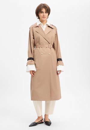Jonge vrouw die een beige trenchcoat met riem draagt, een wit overhemd met verlengde manchetten, lichte broek en zwarte puntige platte schoenen, staande tegen een effen achtergrond.