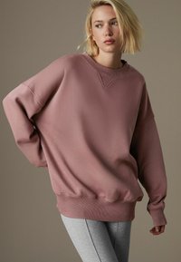 Oversized różowy sweter z okrągłym dekoltem i ściągaczami na rękawach, w połączeniu z szarymi legginsami. Gładki materiał z minimalnymi detalami wizualnymi.