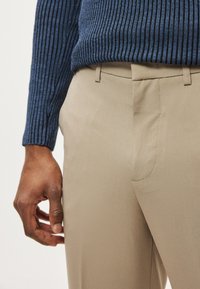 Bézs, testhez álló nadrág sima textúrával és oldalsó slicc zsebekkel, amelyet egy bordázott mintájú, hosszú ujjú tengerészkék pulóverrel párosítanak.