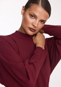 Bordowa sukienka z długim rękawem, o wysokim wycięciu i guzikach na mankietach, wykonana z miękkiego materiału o gładkiej fakturze.