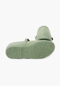 Zapato verde de tela con punta redonda y correa ajustable. La suela de goma texturizada presenta patrones florales y de corazones. Hecho en España.