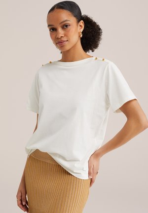 Femeie purtând un top alb cu mâneci scurte cu nasturi aurii pe umeri și o fustă aurie metalică cu striații, pozând cu mâna pe șold.