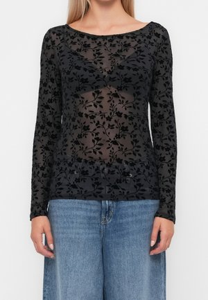 Przezroczysta czarna bluzka z długim rękawem z welurowym kwiatowym wzorem, noszona na czarnym biustonoszu, zestawiona z wysokim stanem niebieskich dżinsów.