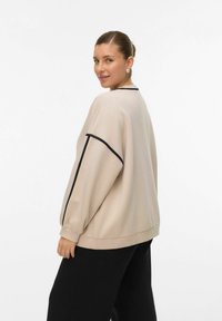 Beige oversized sweatshirt met zwarte piping, ronde halslijn en lange mouwen; gladde textuur; gecombineerd met zwarte broek.