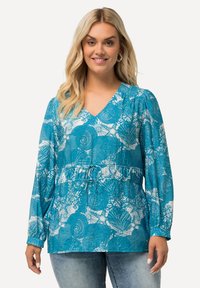 Donna che sorride, indossa una blusa a maniche lunghe color teal con stampa di conchiglie bianche e coralli, abbinata a jeans azzurri, in piedi su uno sfondo bianco.