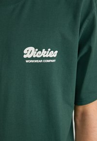 Mørkegrøn bomulds T-shirt med hvid logo tekst "Dickies WORKWEAR COMPANY" på venstre brystområde. Korte ærmer og rund halsudskæring.