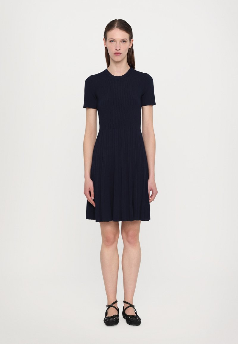 Navy ribbed dress with a short sleeve and round neckline, featuring a flared skirt and pleated design, paired with black ballet flats.