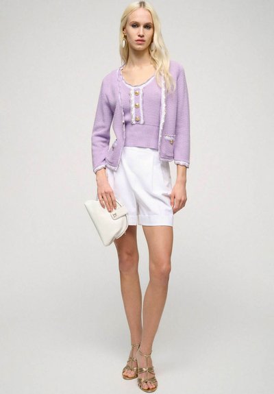Levandulový pletený cardigan s texturovanou tkaninou, ozdobnými knoflíky a bílými detaily, kombinovaný s bílými šortkami a krémovou kabelkou.
