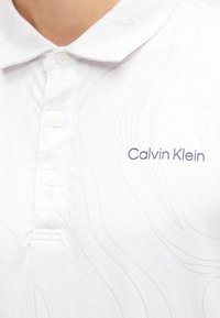Λευκό πόλο πουκάμισο με διακριτικό τοπογραφικό μοτίβο, τρία κουμπιά και το λογότυπο "Calvin Klein" κεντημένο στο στήθος.