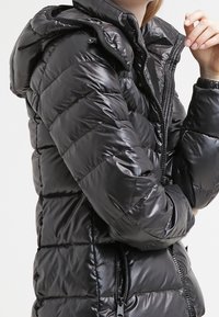 Black, puffy jacket with a glossy finish, featuring a zippered front, hood, and side pockets. Quilted design with visible stitching.