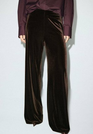 Pantaloni larghi in velluto marrone con una texture liscia, dotati di una vita alta e una leggera lucentezza. Abbinati a una camicia scura.