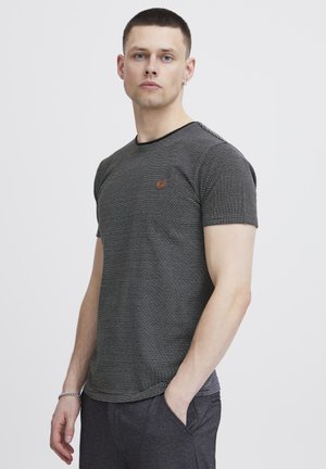 Camiseta de rayas en blanco y negro con cuello redondo, mangas cortas y un pequeño parche de logo marrón en el pecho; textura de tela suave.