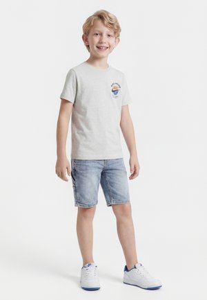 Smilende blond gutt iført en lysegrå Nike-t-skjorte, denimshorts og hvite joggesko med blå såler, stående på en hvit bakgrunn.