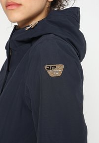 Mörkblå huva jacka med texturerad tyg, med en beige logotyp patch på vänster axel och en dragkedja detalj nära kragen.