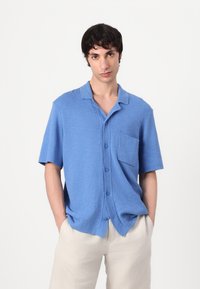 Camisa de botón de manga corta, de color azul, en tejido texturizado con un bolsillo, que presenta un cuello tipo camp y un corte relajado. Se combina con pantalones de color claro.