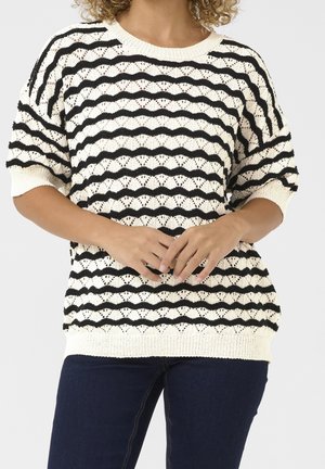 Person trägt einen weißen Strickpullover mit schwarzen wellenförmigen Streifen und halblangen Ärmeln, kombiniert mit dunkelblauen Jeans, die Hände vor dem Körper verschränkt.