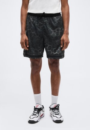 Férfi fekete mintás sportnadrágot, fehér bokazoknit és piros-fekete részletekkel díszített fehér sportcipőt visel, sima háttér előtt áll.