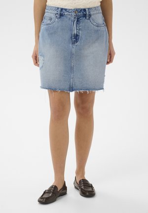 Žena nosi svetlomodrú ošúchanú rifľovú sukňu so strapcami na leme a hnedé mokasíny, stojí na bielom pozadí.