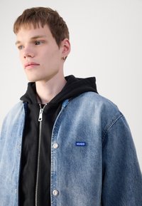 Chaqueta de denim en azul claro con botones de presión plateados y logotipo "HUGO" en azul, superpuesta a una sudadera con cremallera negra. Textura suave, diseño casual.