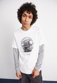 Camiseta blanca con un gráfico de un balón de baloncesto y "Flight" en negro. Largas mangas grises debajo, combinada con pantalones cargo grises.