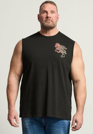 Muskularny mężczyzna w czarnej koszulce bez rękawów z czerwono-szarym emblematem smoka na lewej piersi, stojący na jednolitym tle.